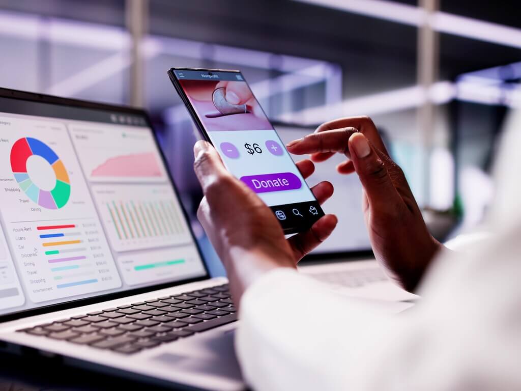 Person using a mobile app to donate money, with financial dashboard visible on laptop.